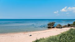 広々としたビーチに小さなカラフルな傘があり、遠くには岩の多い海岸線と青い海が広がり、晴れた夏の日に数人の人が水辺に立っています。