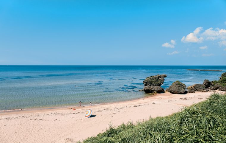 広々としたビーチに小さなカラフルな傘があり、遠くには岩の多い海岸線と青い海が広がり、晴れた夏の日に数人の人が水辺に立っています。