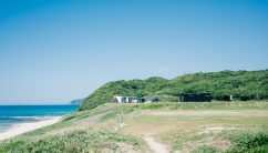 山口県長門市の大浜キャンプ場。青い海と青空が広がる海岸沿いの小高い丘に、いくつかの建物が見える。