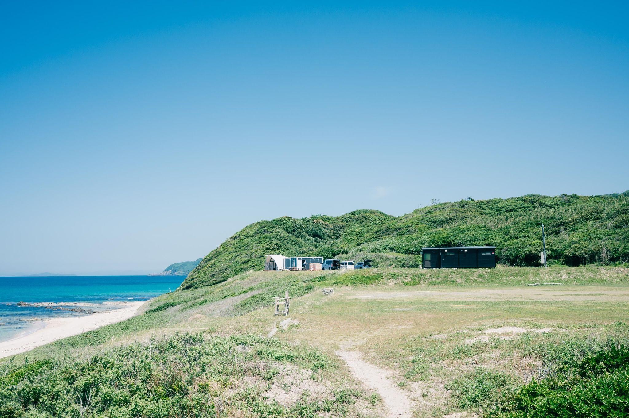 山口県長門市の大浜キャンプ場。青い海と青空が広がる海岸沿いの小高い丘に、いくつかの建物が見える。