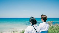 ２人が青い海と空を眺める後ろ姿。風になびく髪と白いシャツが、夏の開放感を演出しています。