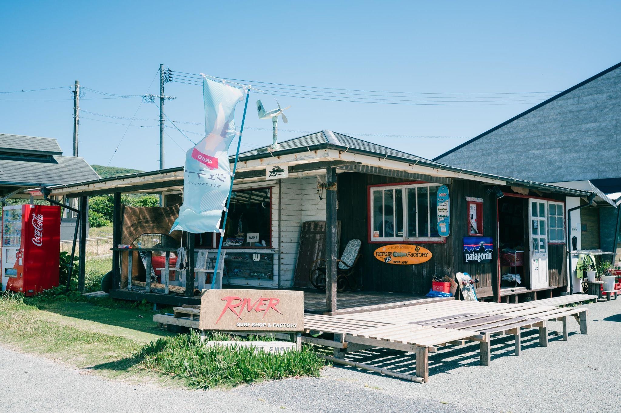 木造の素朴な外観が特徴のRIVER SURF SHOPと書かれたサーフショップ。入り口にはデッキが広がり、サーフィン用品が置かれています。