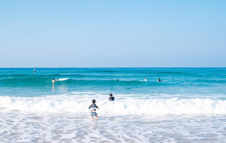 夏の大浜海岸で、子供たちが楽しそうに泳ぐ様子。透き通った青い海に白い波が立ち、夏のレジャーを満喫しています。