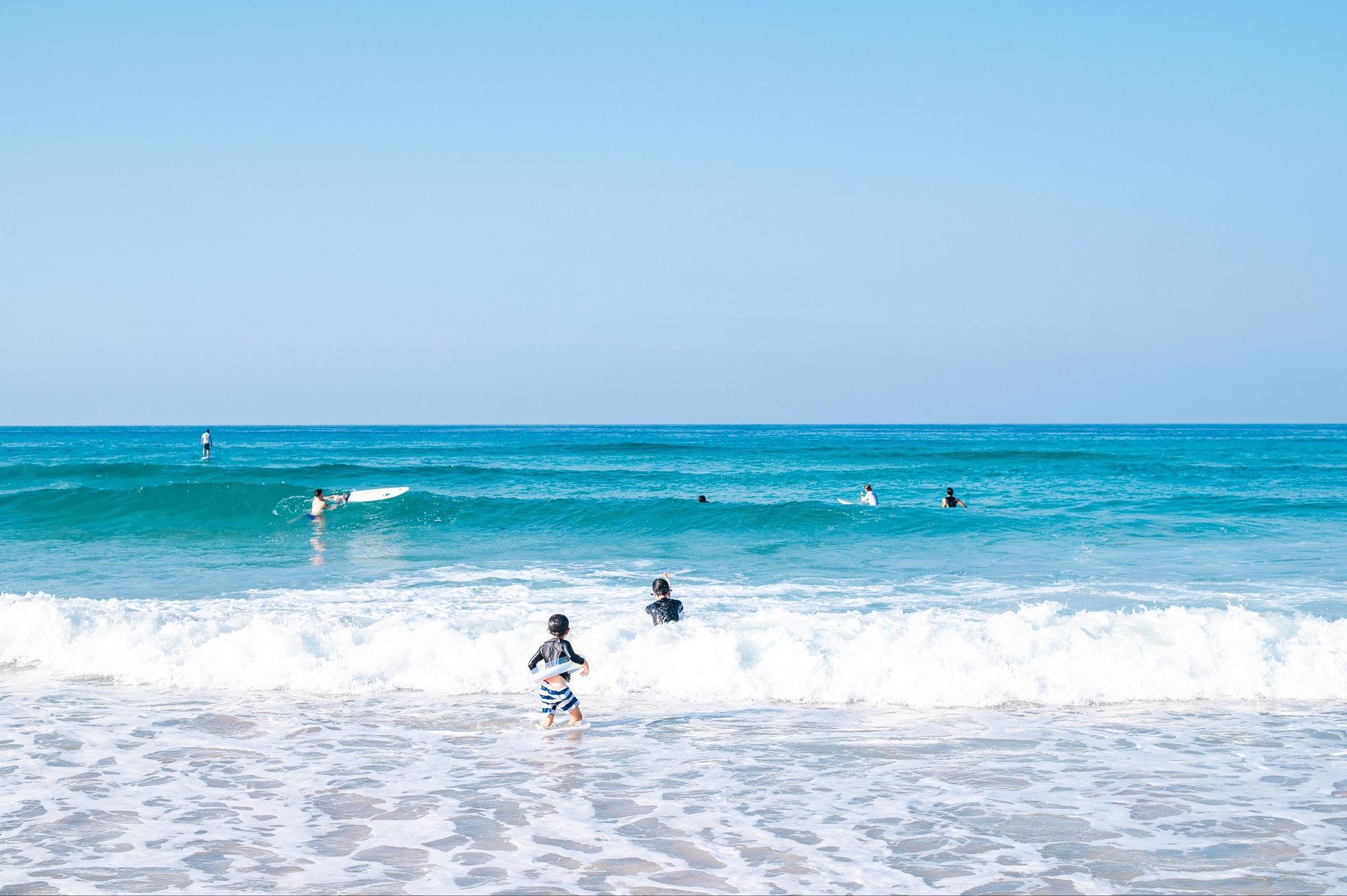 夏の大浜海岸で、子供たちが楽しそうに泳ぐ様子。透き通った青い海に白い波が立ち、夏のレジャーを満喫しています。