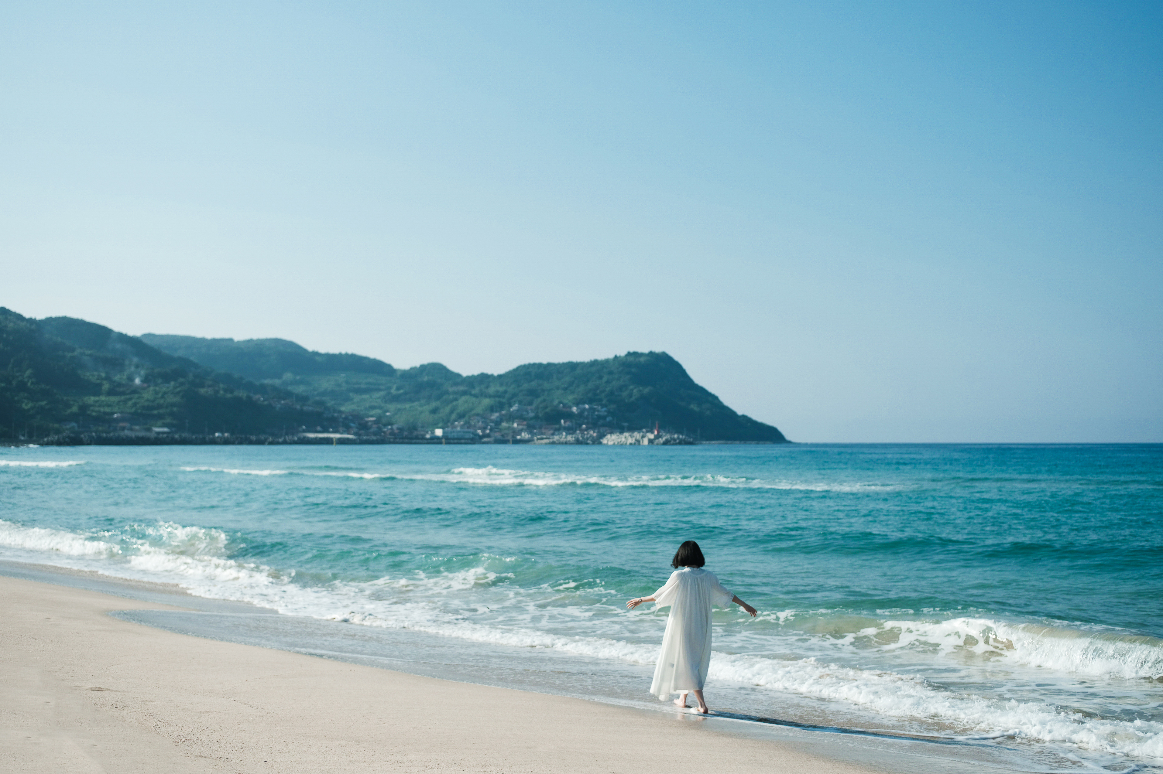 白いドレスを着た女性が、青い海と山々を背景にした美しい砂浜の波打ち際を歩いている。