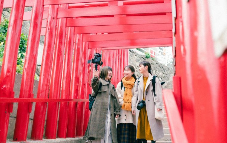 元乃隅神社の朱色の鳥居が続く階段を、ビデオカメラを持つ女性を含む3人の友人が楽しそうに歩いています。