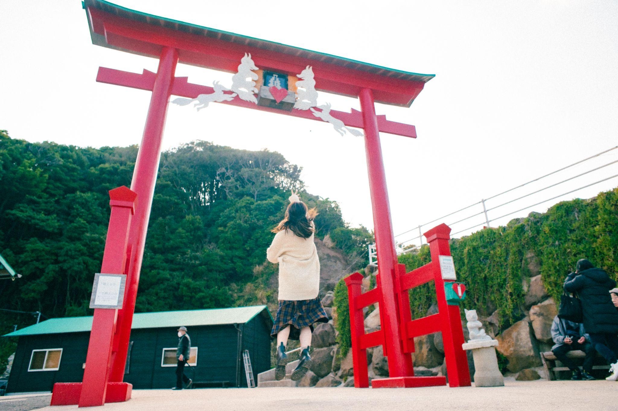 元乃隅神社の巨大な朱色の鳥居の前で、女性が空に向かって手を伸ばし、楽しんでいる様子です。
