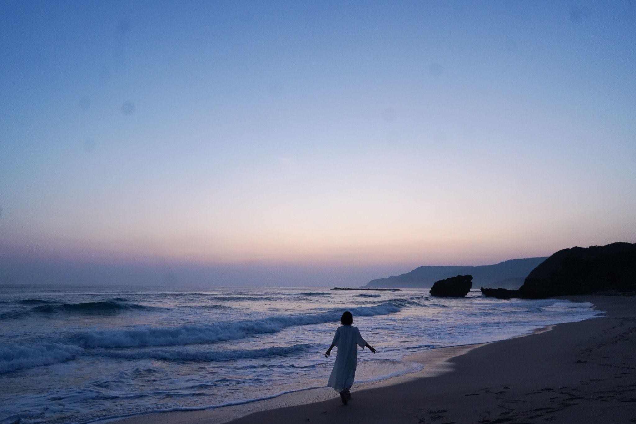 夕暮れ時の砂浜を歩く白いドレスの女性。遠くに山々が見え、穏やかな波が打ち寄せている。